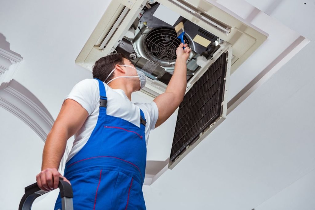 A technician in a mask services a ceiling air conditioning unit, using a brush to clean its components.