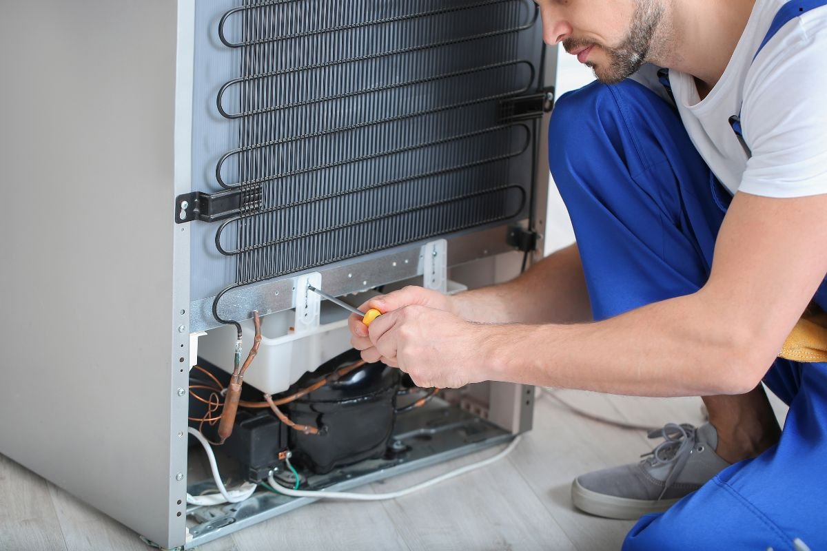 A technician in blue overalls repairs a refrigerator's inner mechanics with a screwdriver, focusing on the condenser coils.