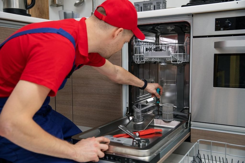 A technician in a red shirt and cap repairs a dishwasher, using tools while inspecting its interior.
